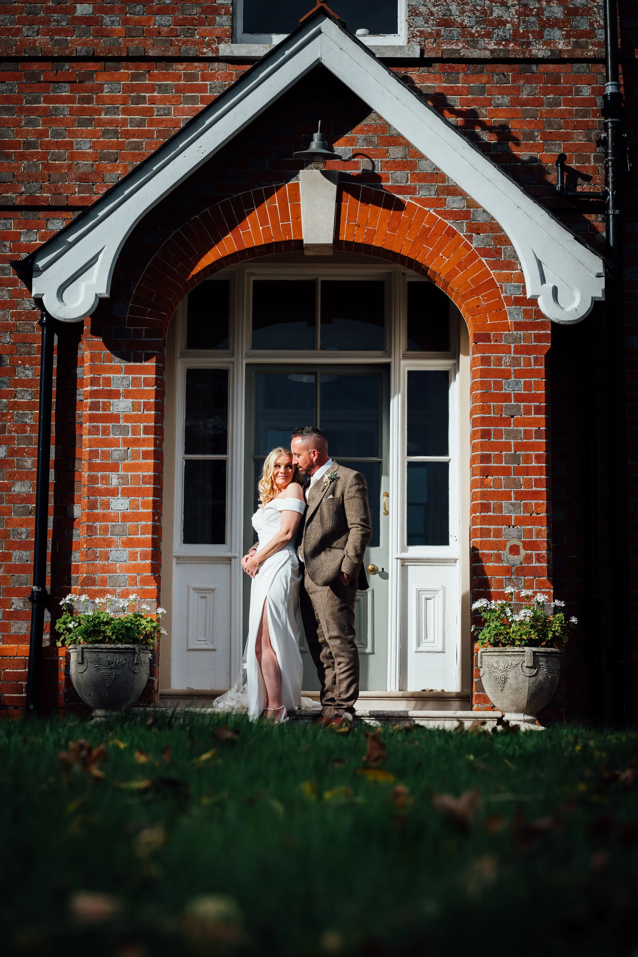 A couple dressed in wedding attire standing close together in front of a brick house with a white door and windows, surrounded by two large potted plants with flowers.