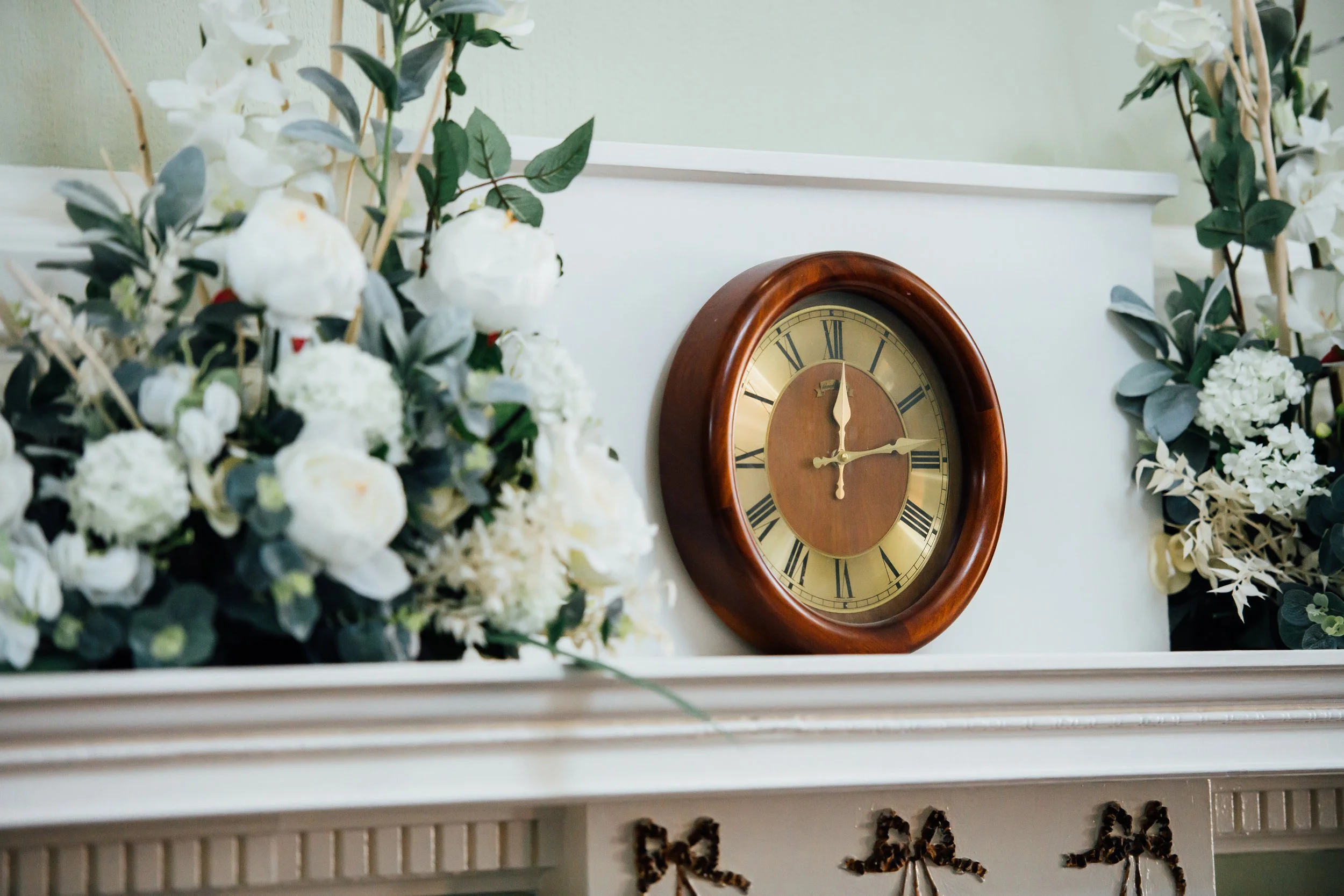 A decorative clock with Roman numerals and a wooden frame hanging above a fireplace mantel decorated with white flowers and greenery.