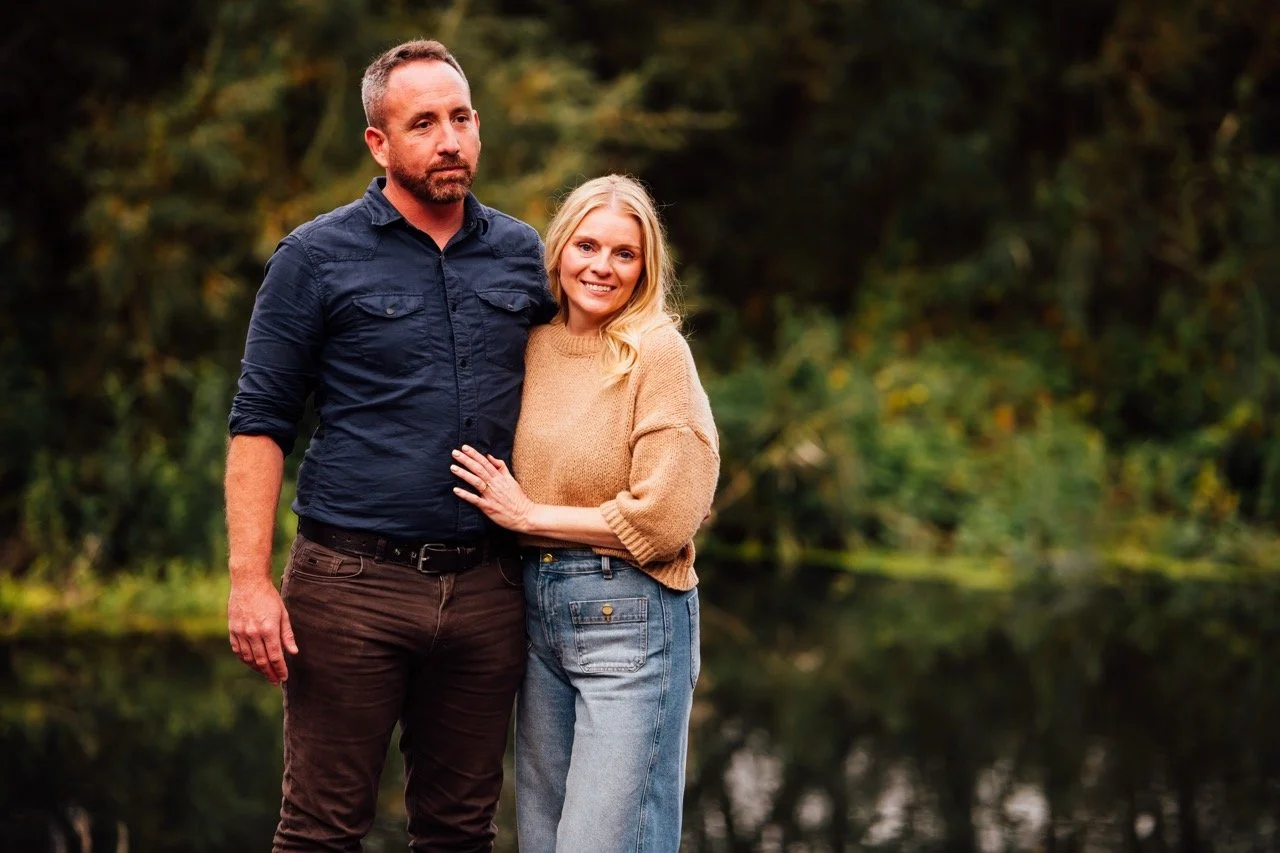 A man and woman standing outdoors near water with green trees in the background, smiling and posing for a photo.