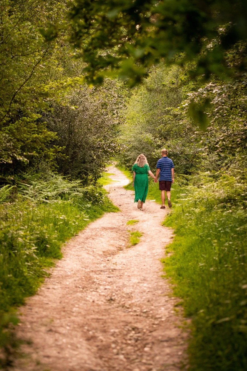 A couple holding hands walking down a dirt path surrounded by lush green trees and foliage.