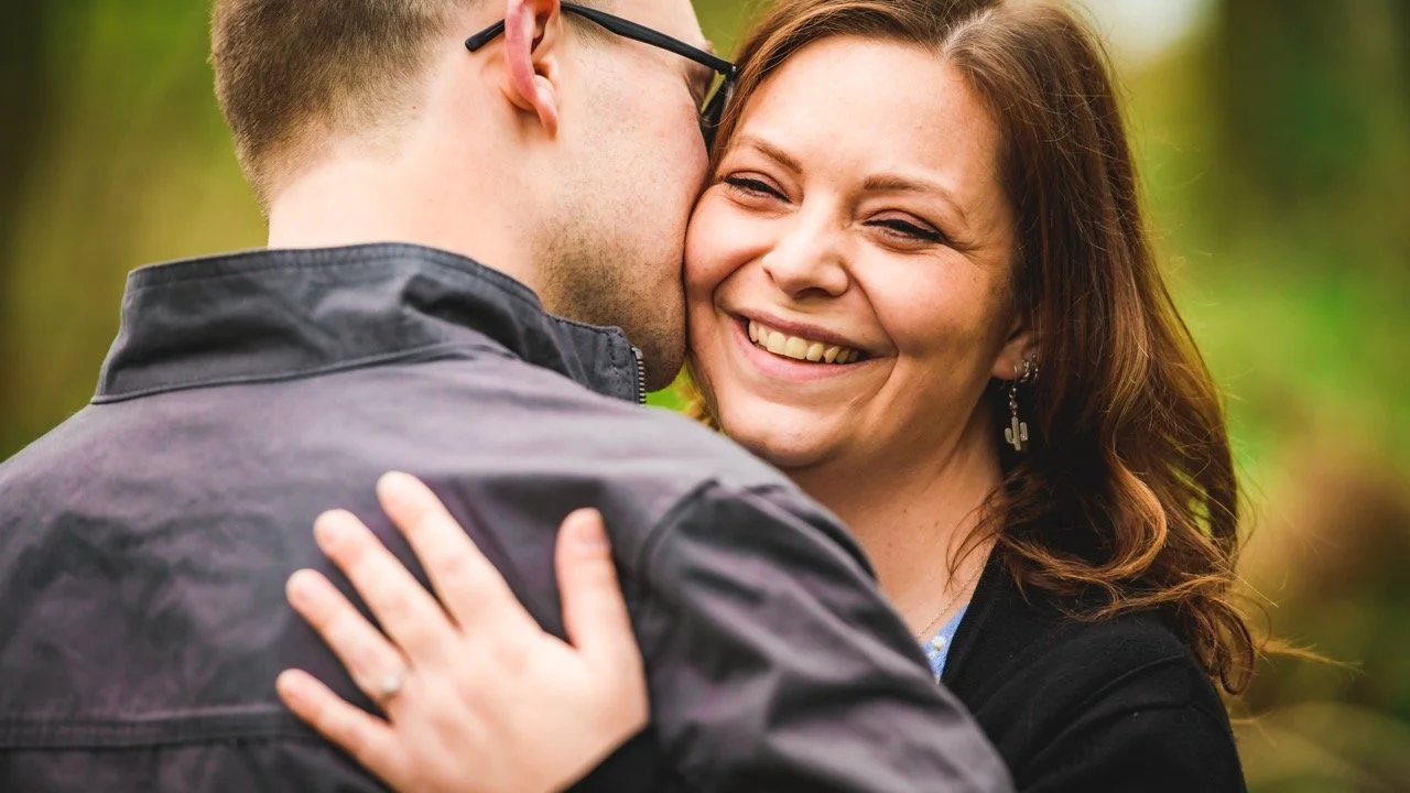A man and woman sharing a close embrace outdoors, the woman smiling happily as she looks at the man.