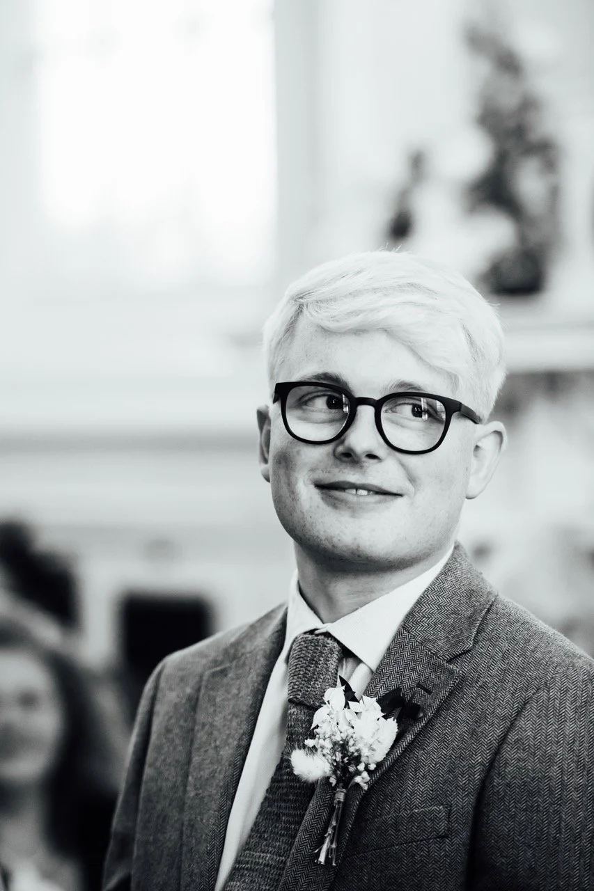 A young man with blond hair and glasses wearing a suit and tie, with a floral boutonniere, smiling in a black and white photo, in an indoor setting.