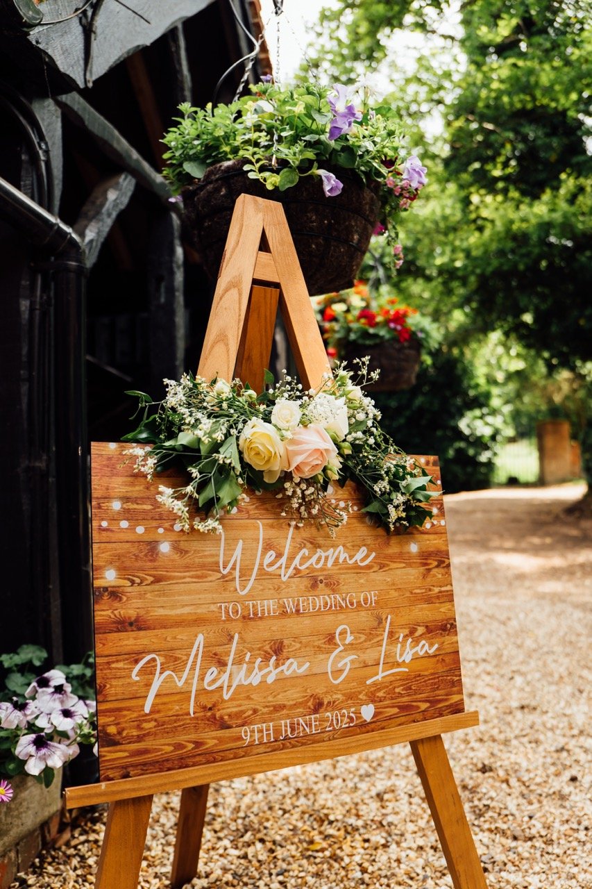 A wooden wedding welcome sign decorated with white and blush roses and greenery, placed on an easel outside, with potted flowers and trees in the background.