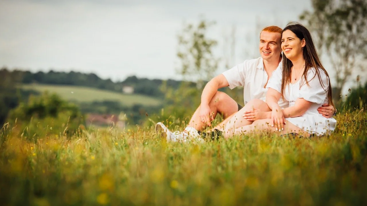 A young couple sitting on the grass in a field, smiling and enjoying the outdoors on a bright day.