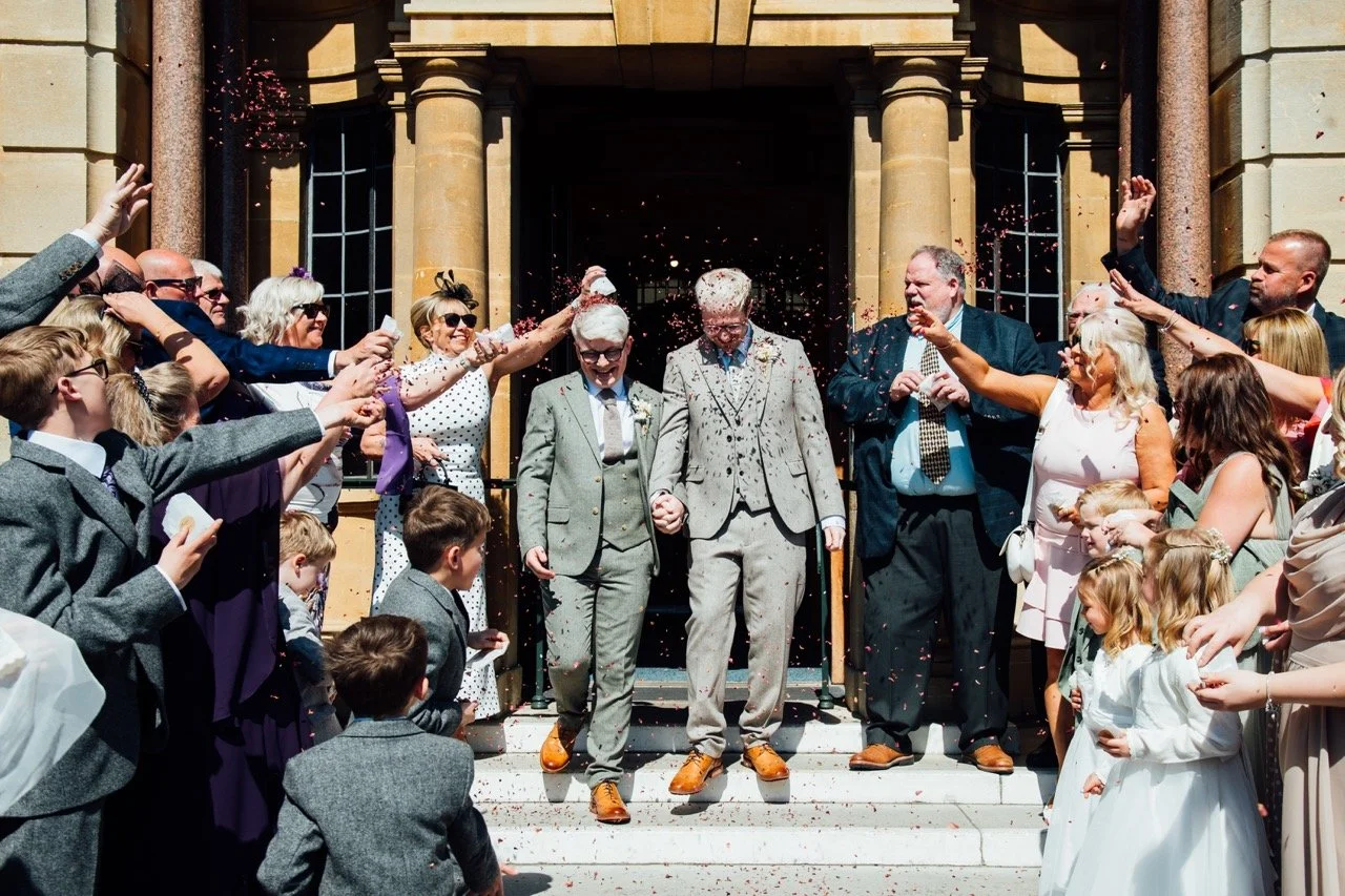 Gay couple walking down the steps of a church while being showered with confetti by family and friends after their wedding ceremony.