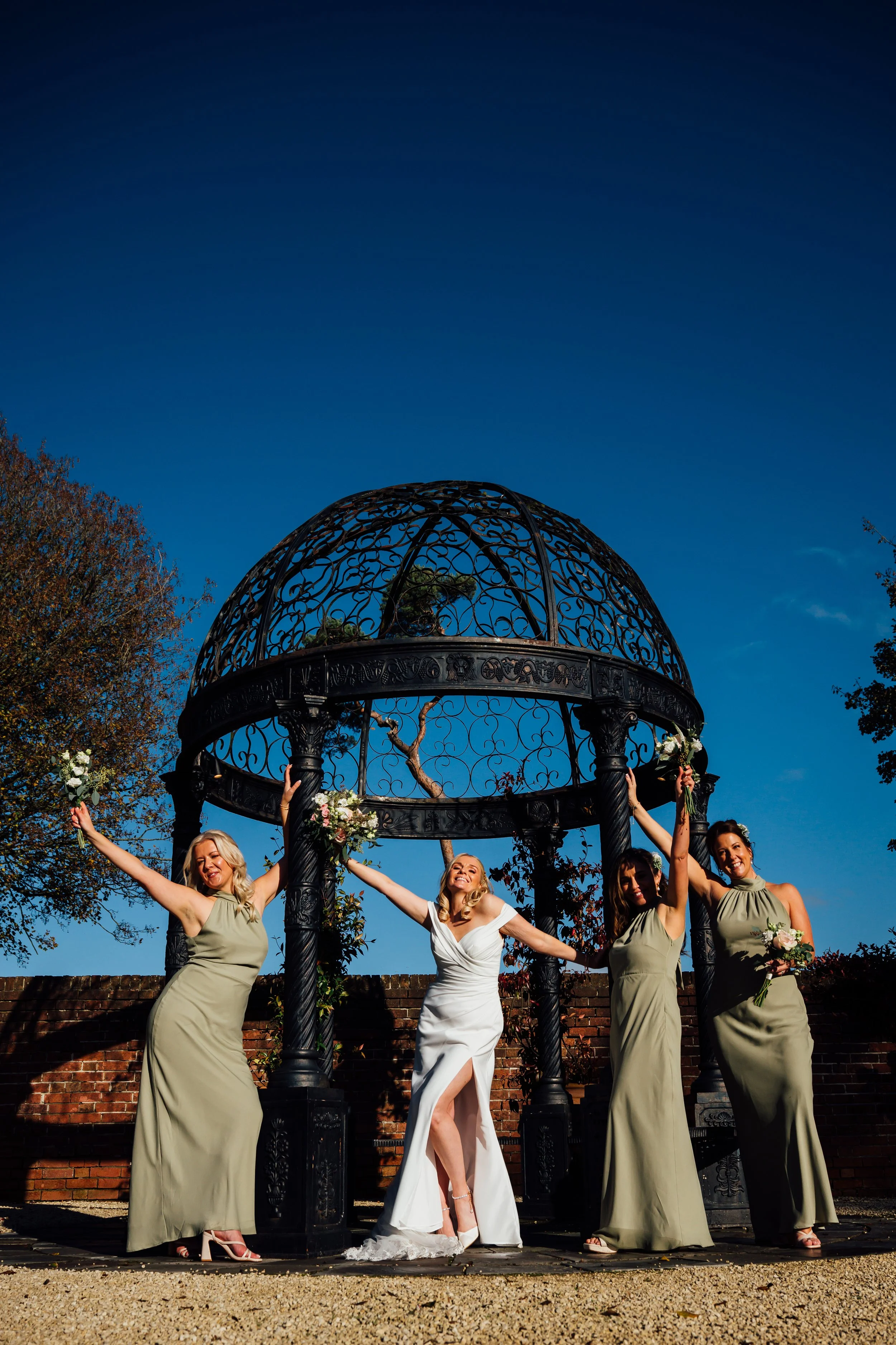 Four women in evening dresses celebrating under a decorative black wrought iron gazebo in a garden on a sunny day.