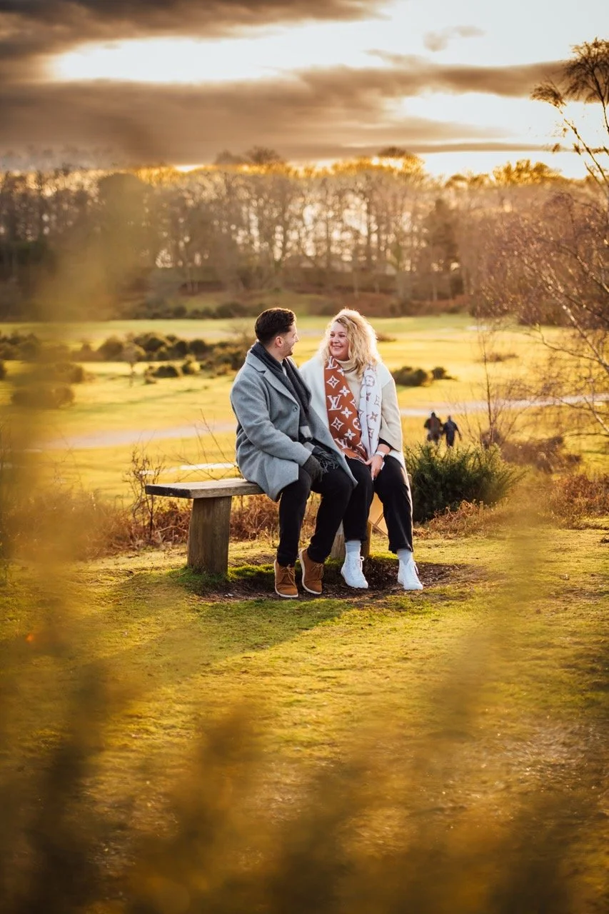 A young couple sitting on a wooden bench in a park during sunset, smiling and looking at each other with a scenic landscape of trees and open fields in the background.