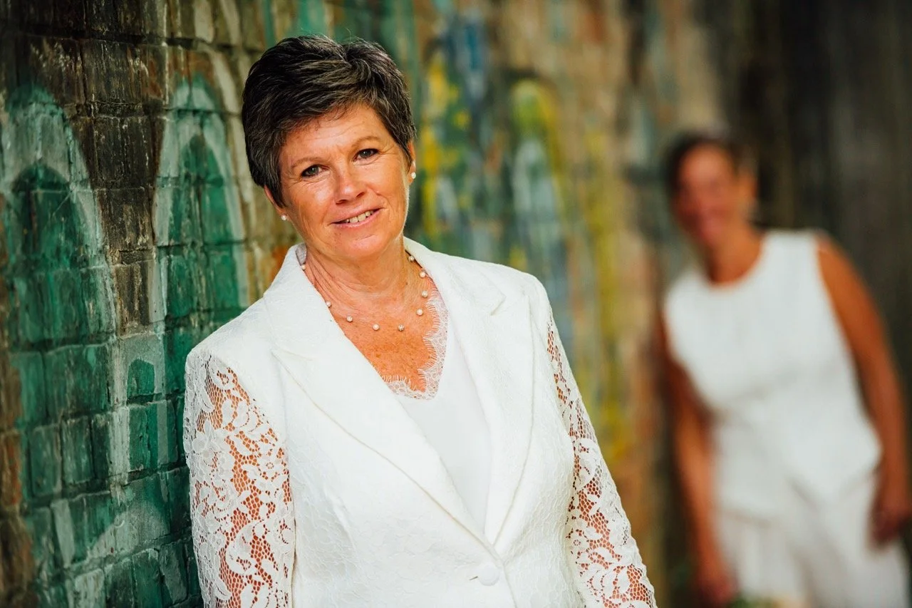 A woman with short dark hair wearing a white lace blazer and necklace standing against a colorful brick wall, with another woman in a white dress blurred in the background.