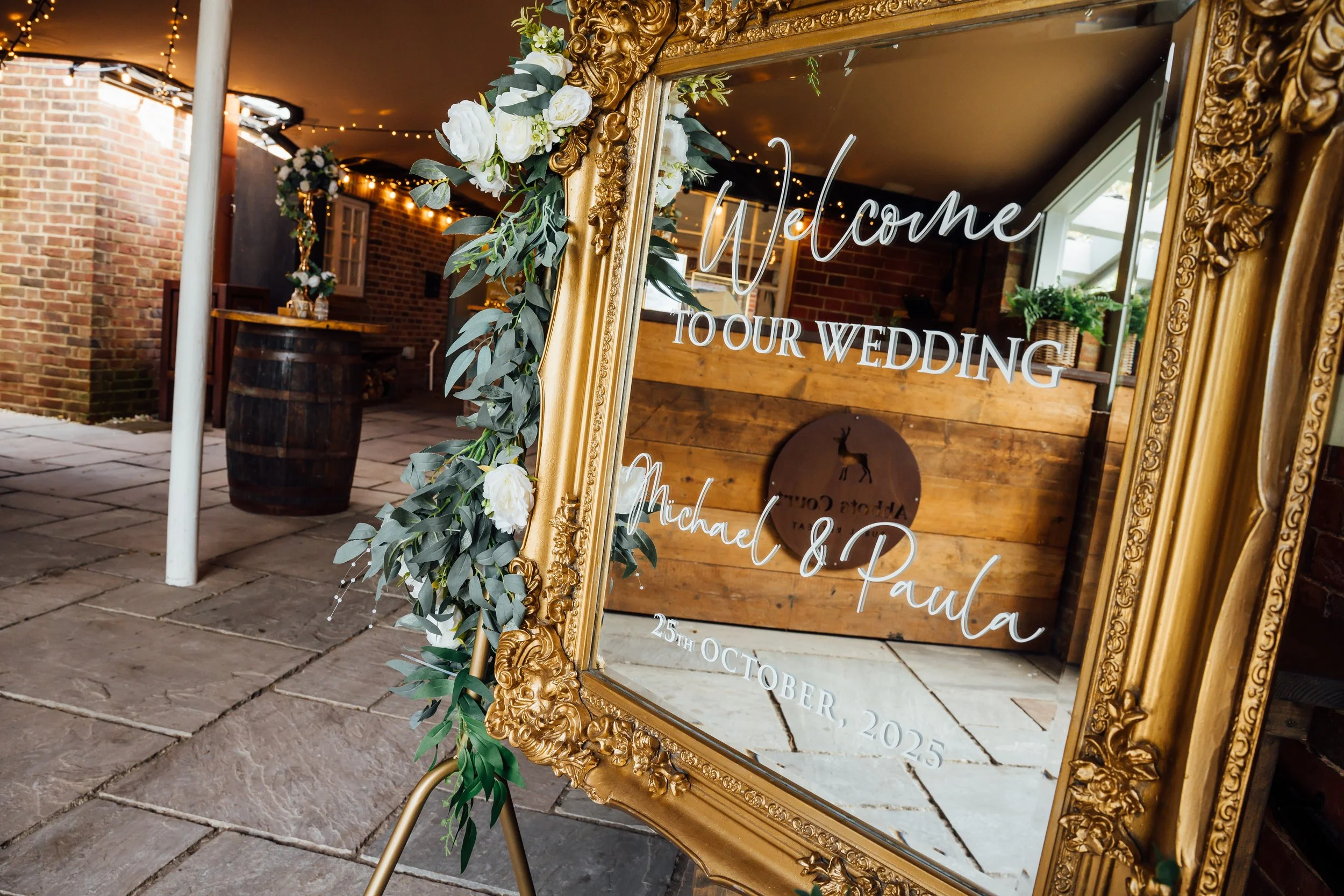 Decorative mirror with gold frame adorned with flowers, displaying a welcome message for a wedding, with printed text including the couple's names, Michael and Paula, and the wedding date, October 25th, 2025.