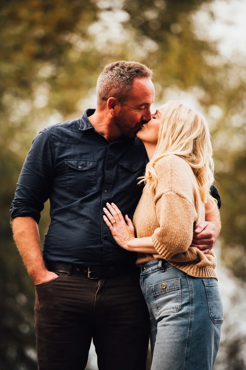 A man and woman sharing a kiss outdoors, with trees and blurred natural background.