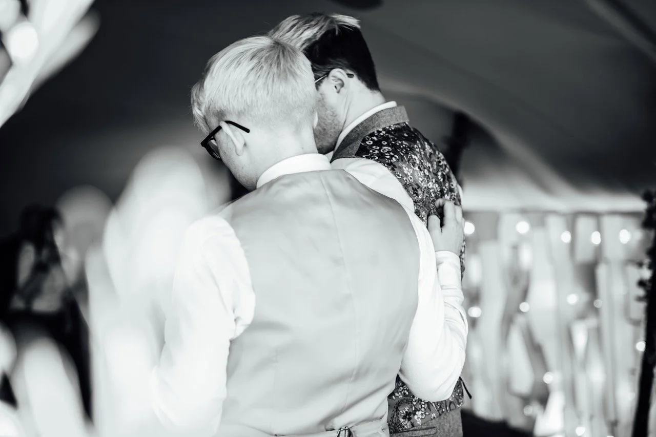 Two men embracing at a celebration or wedding, standing closely together with eyes closed.