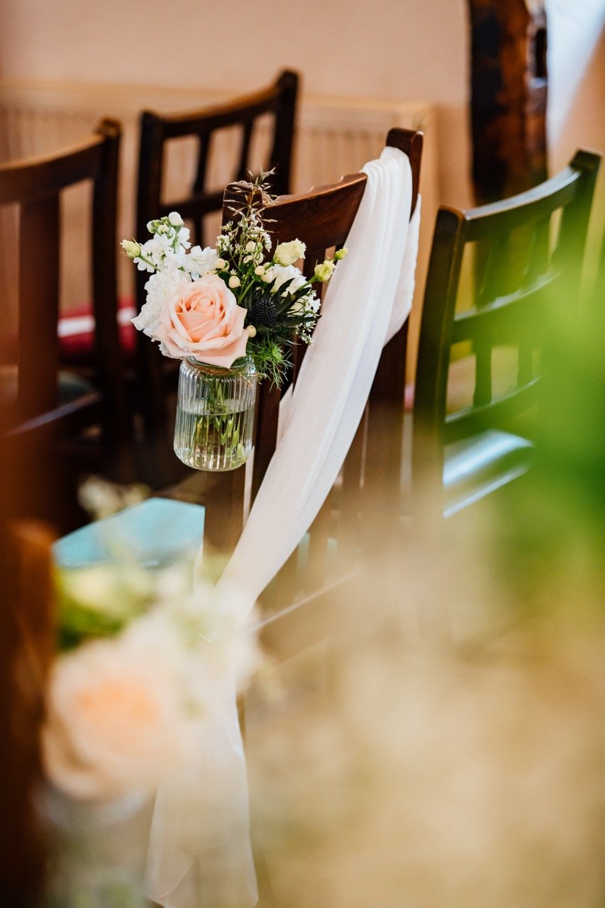 A decorative flower arrangement hanging from the back of a brown wooden chair in a room with similar chairs, white fabric draped over the chair, and blurred elements in the foreground and background.