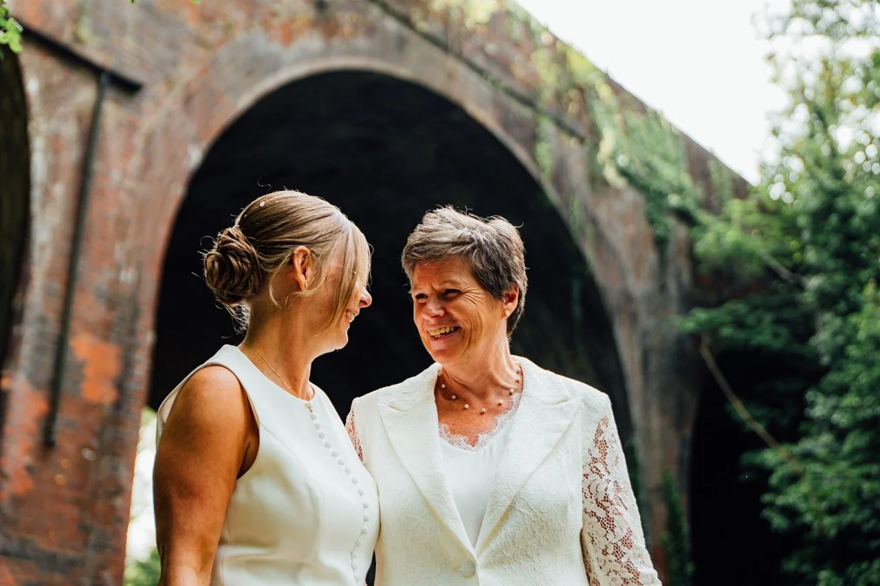 Two women in white dresses smiling and facing each other outdoors near a brick tunnel surrounded by greenery.
