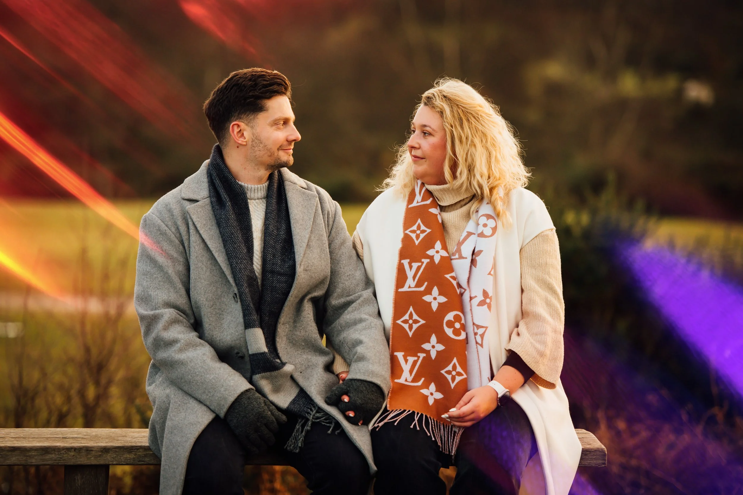 A man and woman sitting next to each other on a wooden bench outdoors during fall, looking at each other with a blurred autumn background.