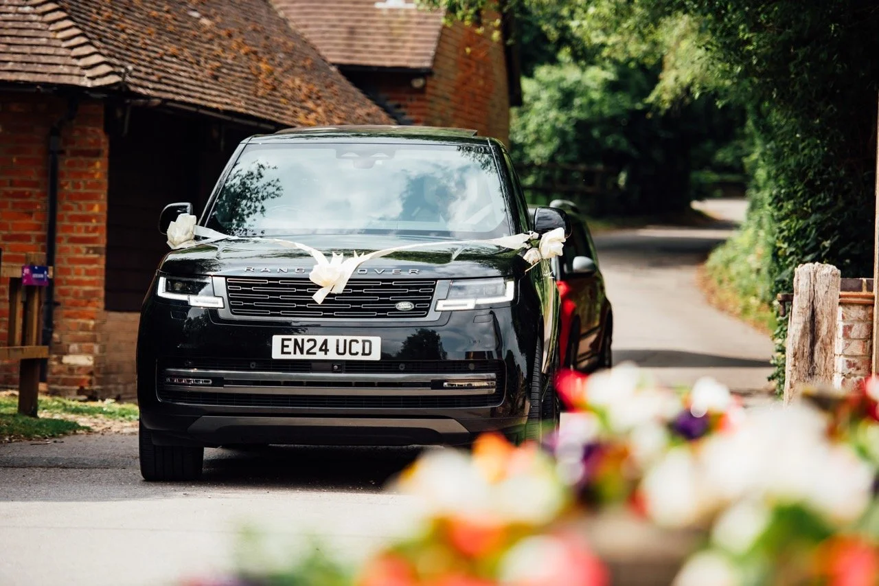 A black Range Rover decorated with white ribbons for a wedding, parked on a residential street with greenery and brick houses in the background.