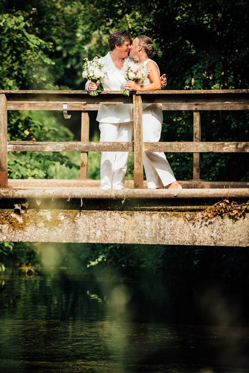Two women, dressed in white, standing on a wooden bridge with green trees in the background, sharing a kiss and holding bouquets of flowers.