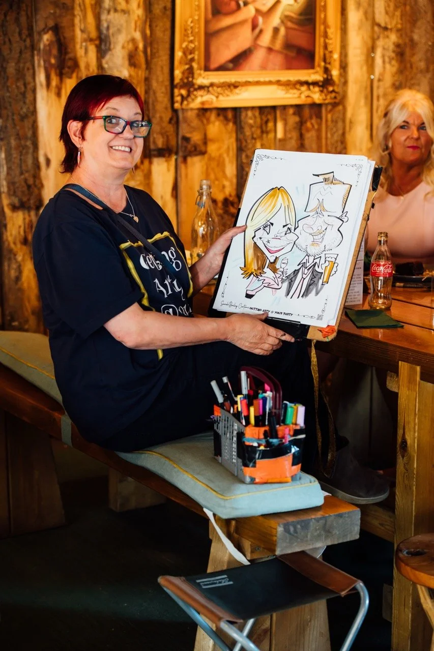 A woman with short dark red hair and glasses holds a caricature drawing of herself and another woman. She is smiling and sitting on a wooden bench at a rustic indoor venue with wood-paneled walls. There are drinks and art supplies on the table in fro