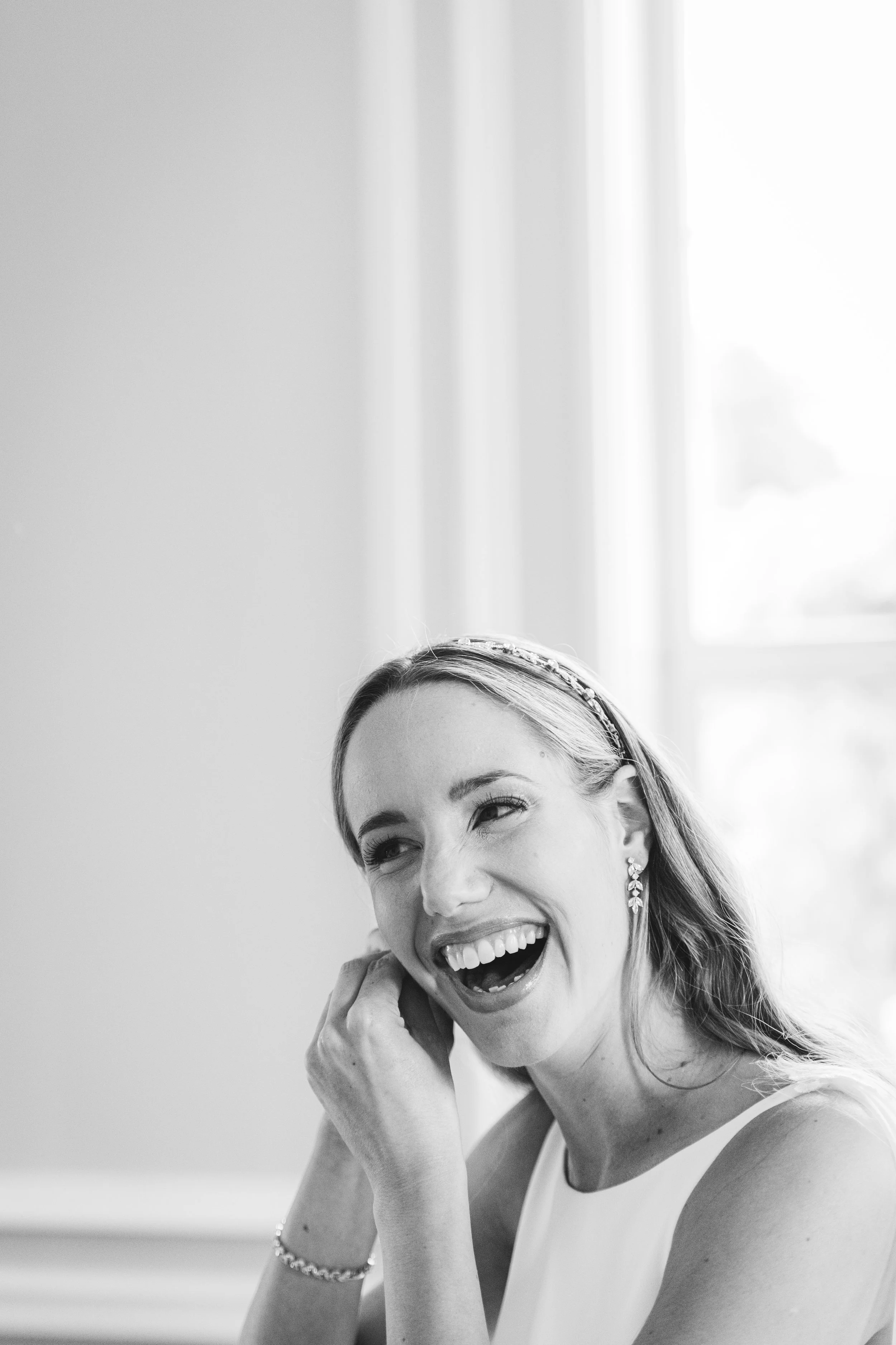 A woman smiling and laughing, wearing earrings, a headband, and a sleeveless top, indoors with a window and curtains in the background.