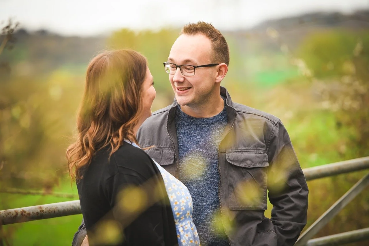 A man and woman smiling and talking outdoors in a park, with blurred trees and greenery in the background.