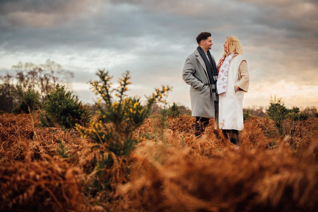 A man and woman standing in a field of brown plants and bushes, smiling at each other under a cloudy sky during sunset.