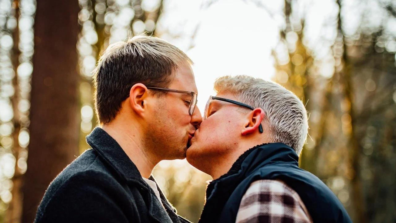 Two men with glasses kiss outdoors in a wooded area during sunset.