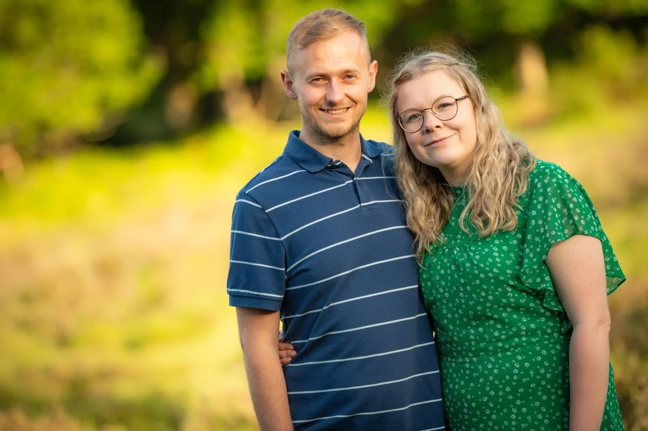 A man and woman standing outdoors in a natural setting, smiling at the camera, with green trees in the background.