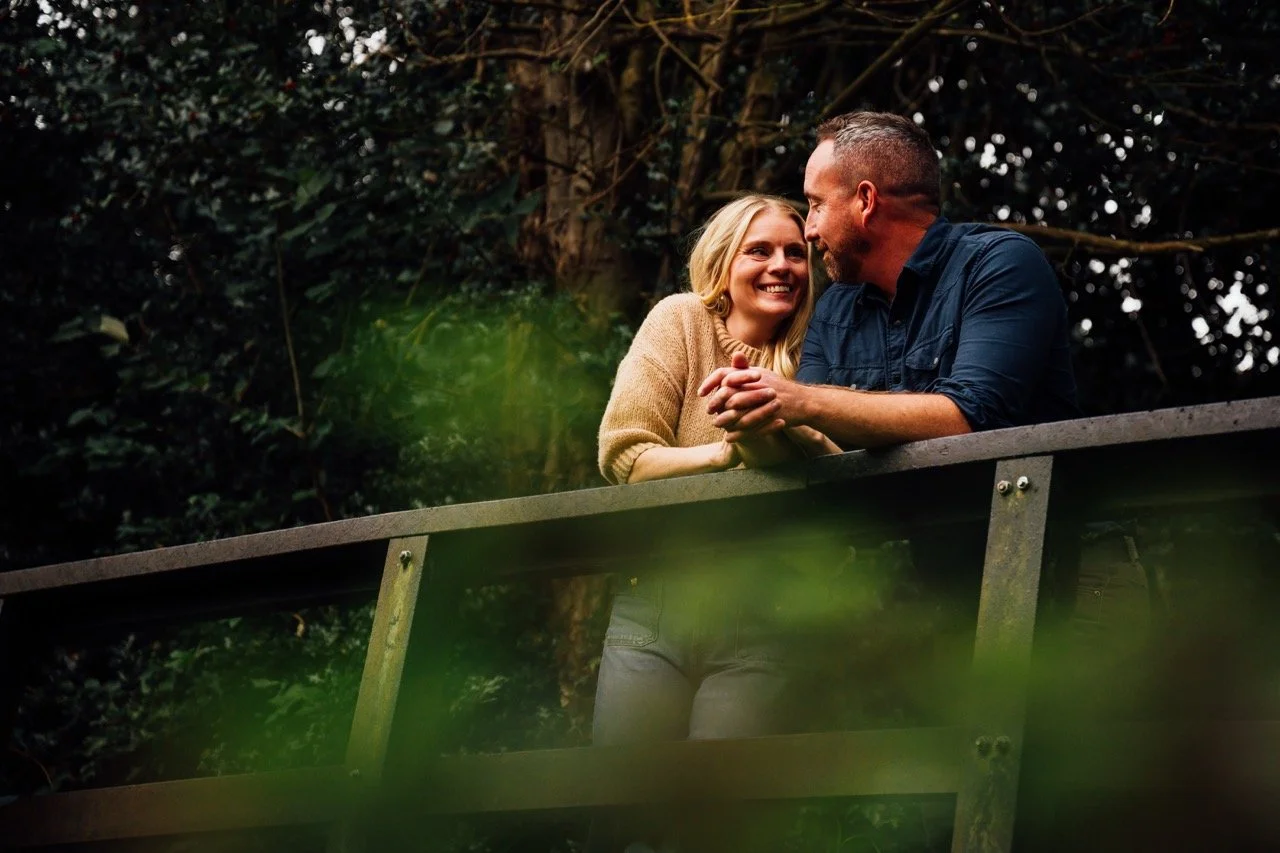 A couple standing on a wooden bridge, smiling and holding hands, surrounded by lush greenery and trees.