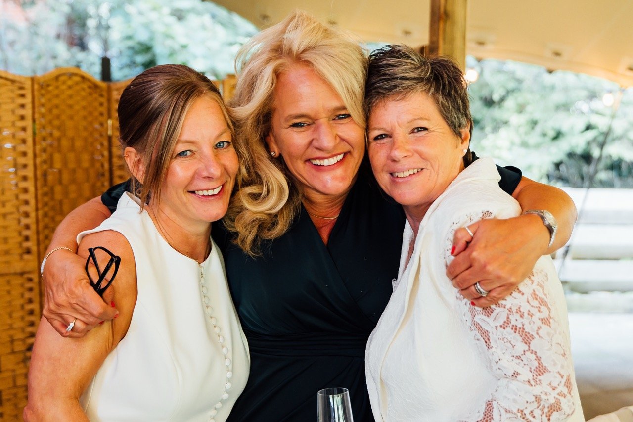 Three women hugging and smiling at an outdoor gathering.