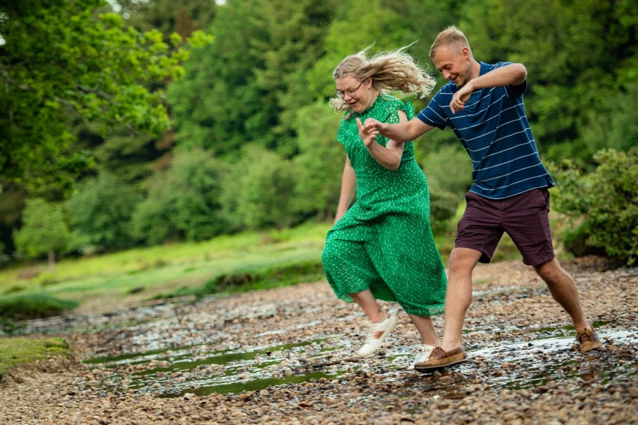 A middle-aged woman in a green dress and a young man in a striped shirt are holding hands and laughing while walking through a shallow creek in a green outdoor park.