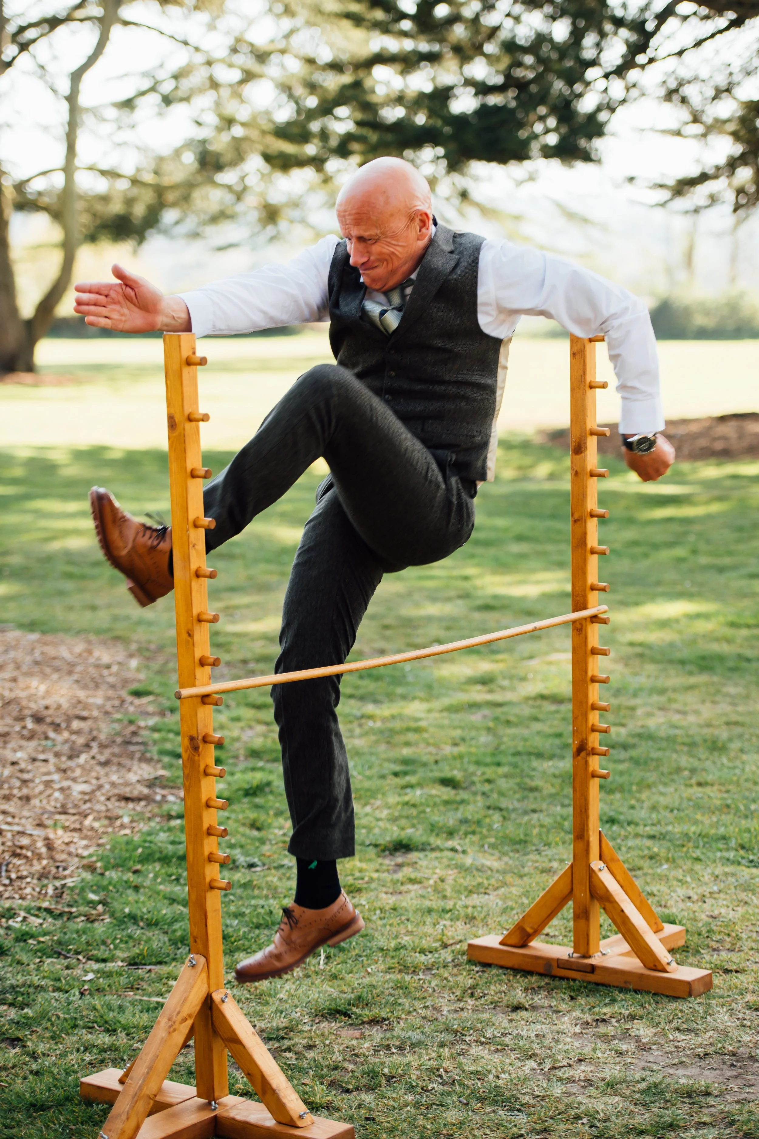 A bald man in a suit is jumping over a wooden agility obstacle outdoors in a park.