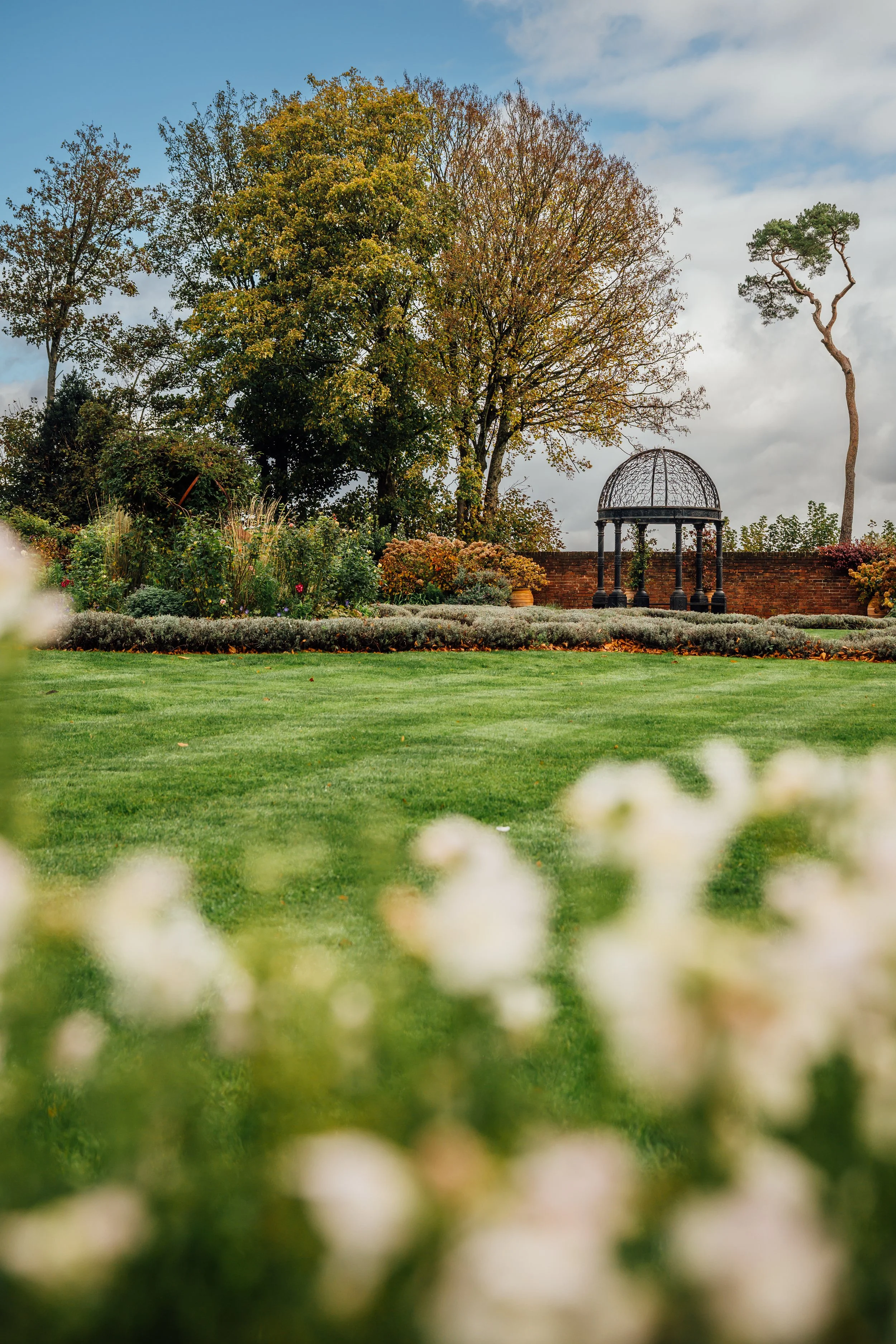 A landscaped garden with a well-maintained grass lawn, bushes, flowers, and tall trees, including one with a curved trunk, under a partly cloudy sky. There is also a structure with a dome-shaped top and columns, enclosed by a brick wall.