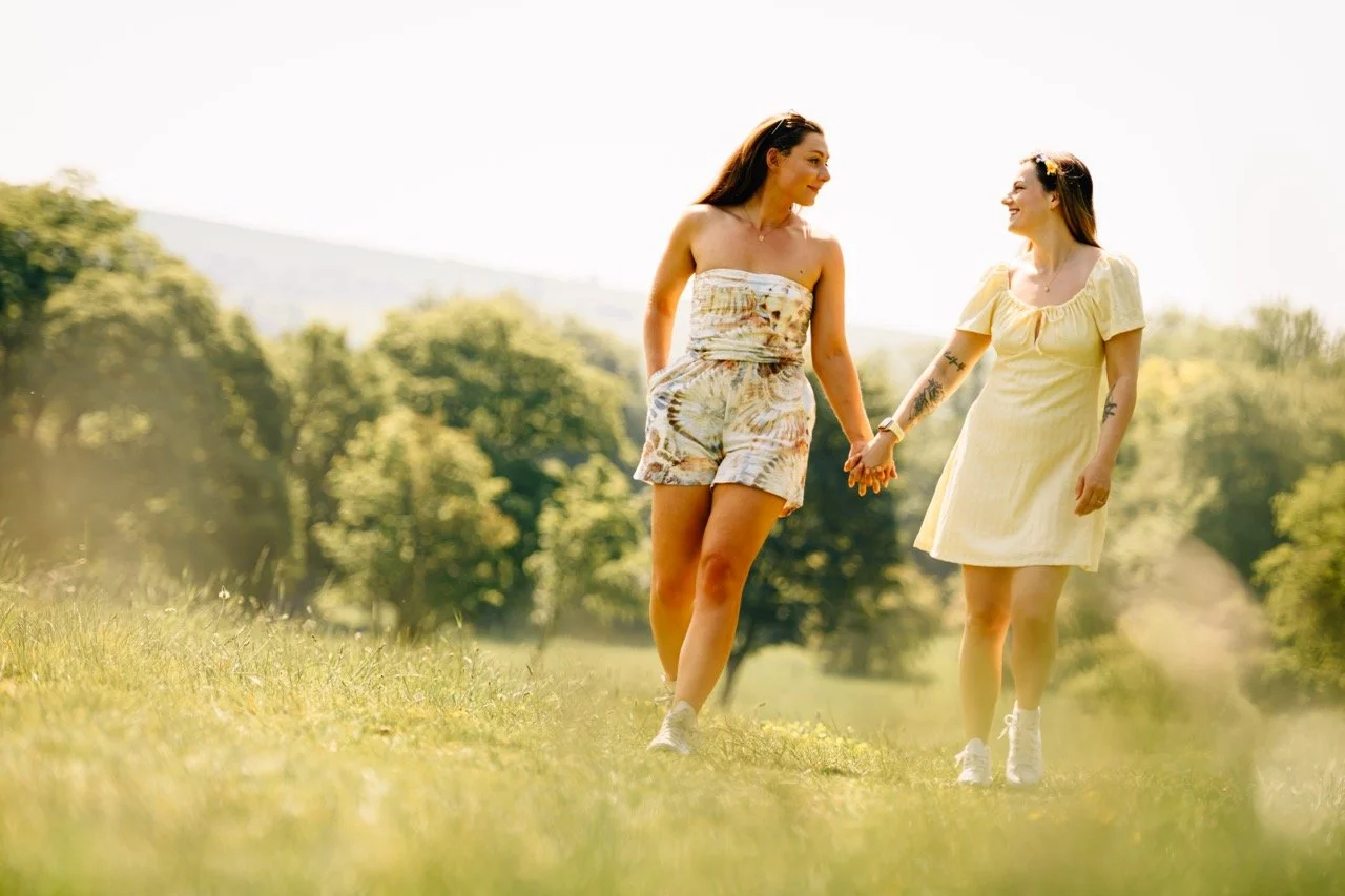 Two women holding hands and walking through a grassy park on a sunny day, smiling and enjoying each other's company.