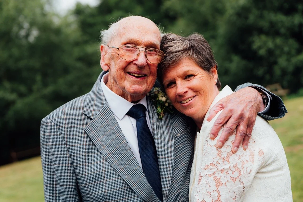 An elderly man and a woman smiling and hugging outdoors in a park or garden setting.