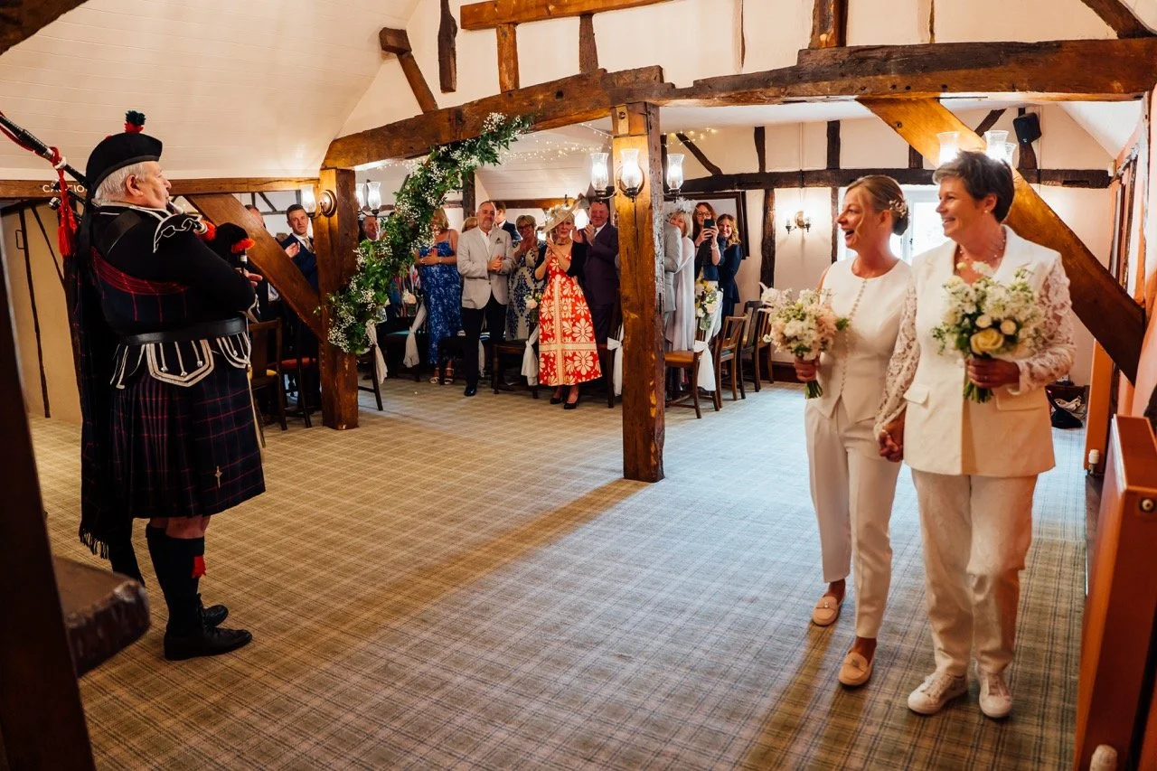 A wedding ceremony with two women in white holding bouquets, walking towards a man in traditional Scottish attire, in a decorated rustic venue with guests watching.