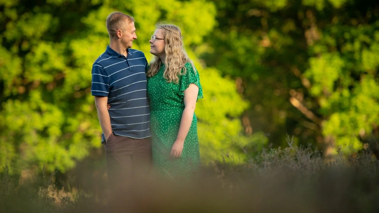 A man and woman standing together outdoors, looking at each other in a green, wooded area.