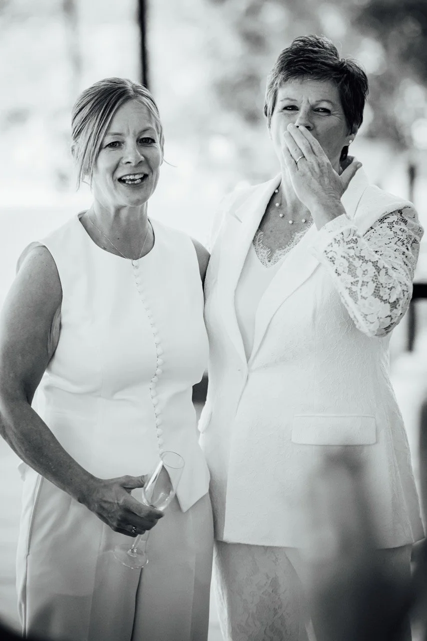 Two women in white dresses at a social gathering, one holding a wine glass, the other appears to be mid-laugh covering her mouth.