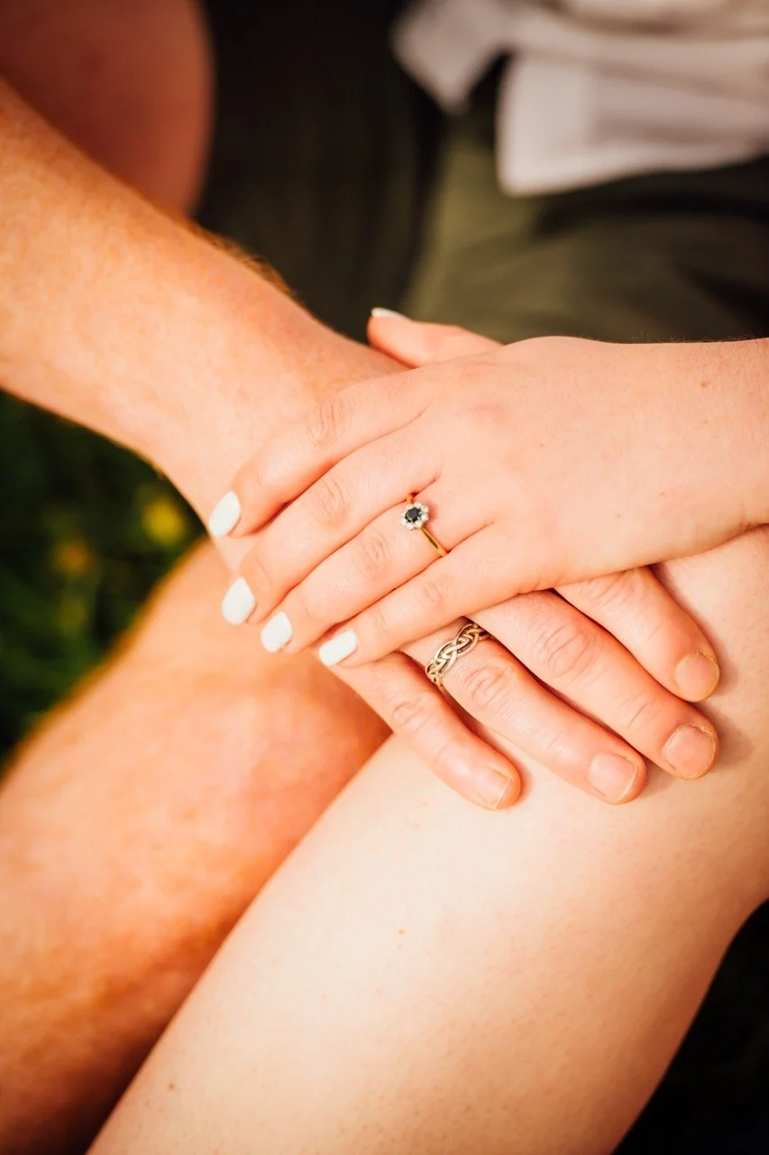 Close-up of a couple holding hands, showing their wedding rings, with the focus on the woman's hand with a black gemstone ring and white nail polish.