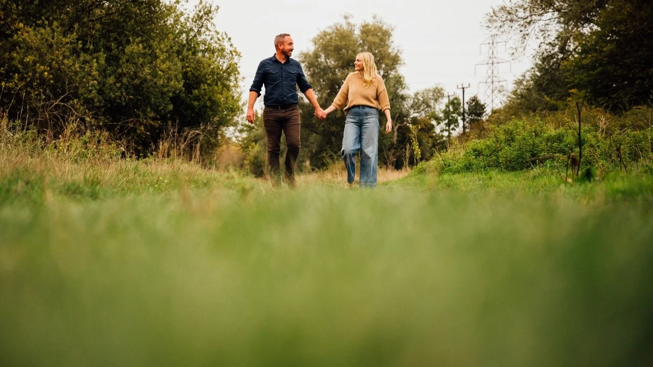 A couple holding hands and walking through a grassy field surrounded by trees during daytime.