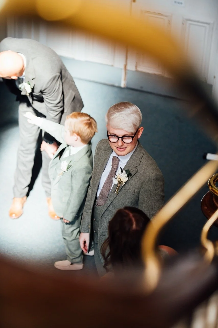 A group of people, including an older man with gray hair and glasses and two young boys, are seen through a circular frame, likely taken from a staircase or balcony. They are dressed in formal attire, possibly at a wedding or special event.