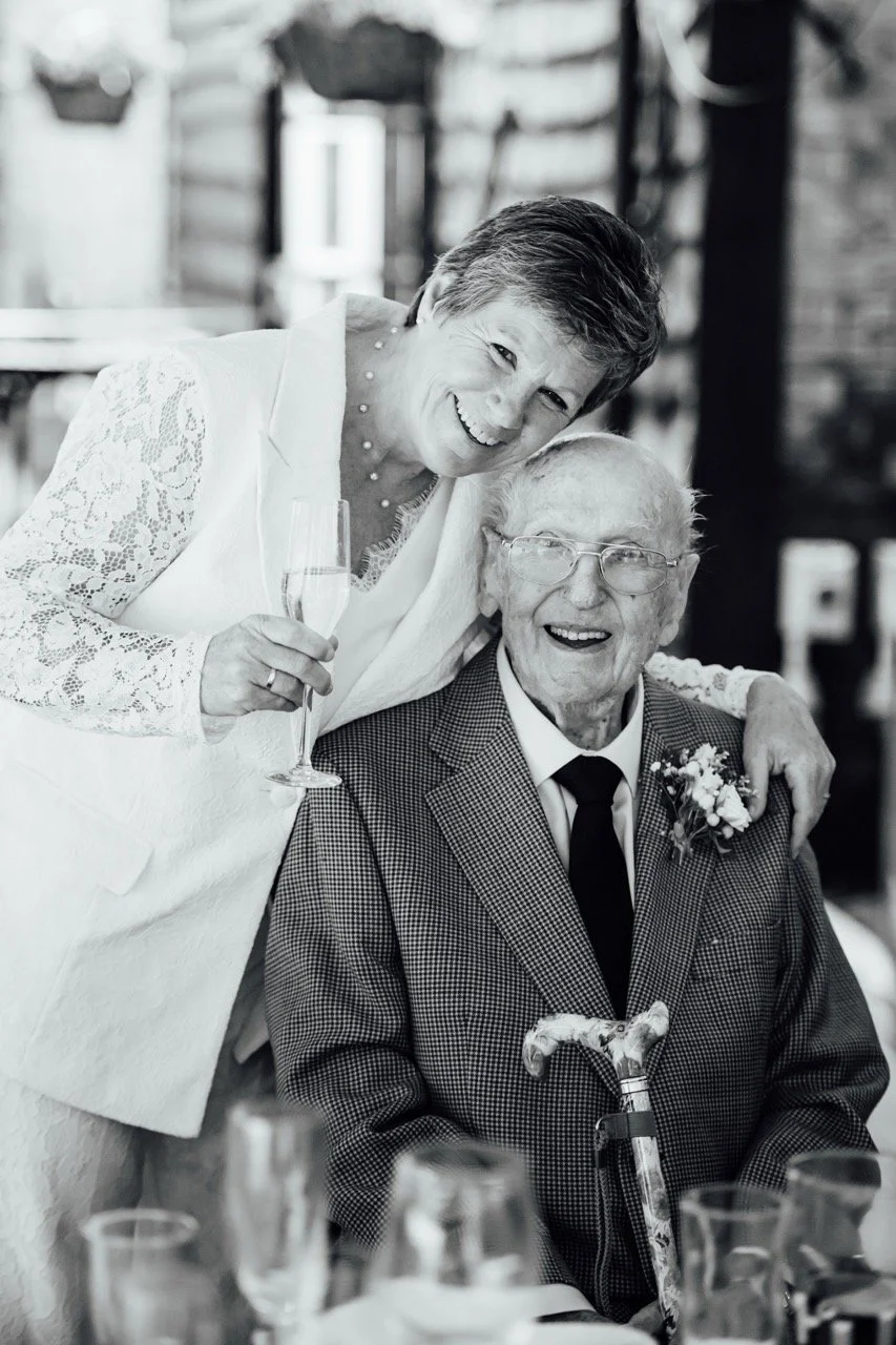 A smiling elderly man with glasses and a cane, wearing a suit with a boutonniere, seated at a table, and a woman with short hair and a lace dress, happily leaning over him with a glass of champagne in her hand, in a festive setting.
