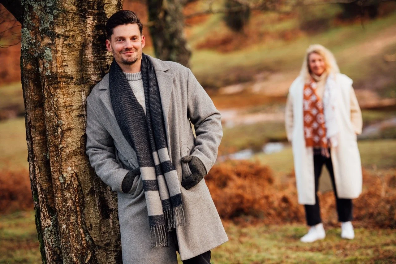 A man with dark hair and a beard leans against a tree, smiling, in a park during fall. He wears a gray coat, a gray and black striped scarf, and gloves. In the background, a woman with blonde hair, dressed in a white coat and orange patterned shirt, 