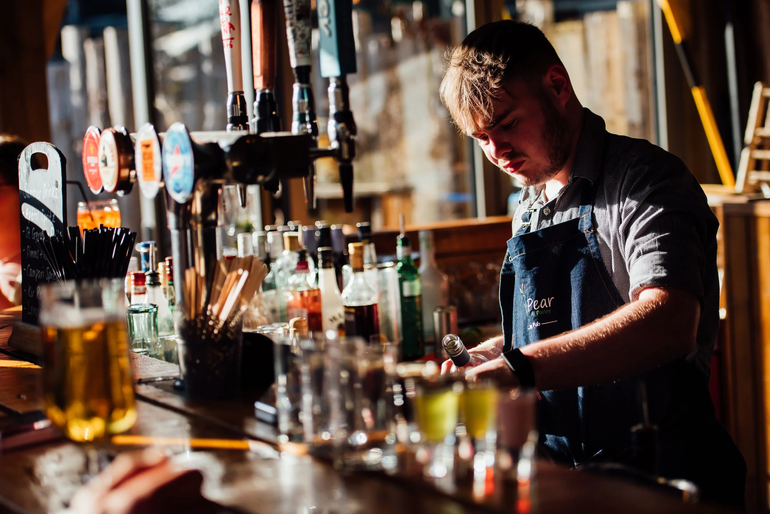 A young man working behind the bar at a pub or restaurant, preparing a drink, with various bottles and bar tools on the counter, illuminated by natural light from windows.