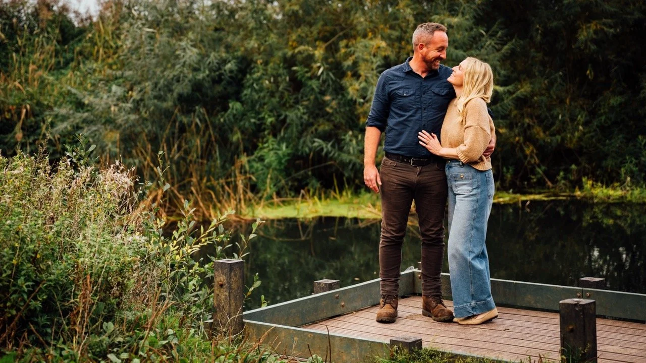 A couple standing on a wooden dock near a pond, embracing and smiling at each other in a natural outdoor setting with lush greenery in the background.