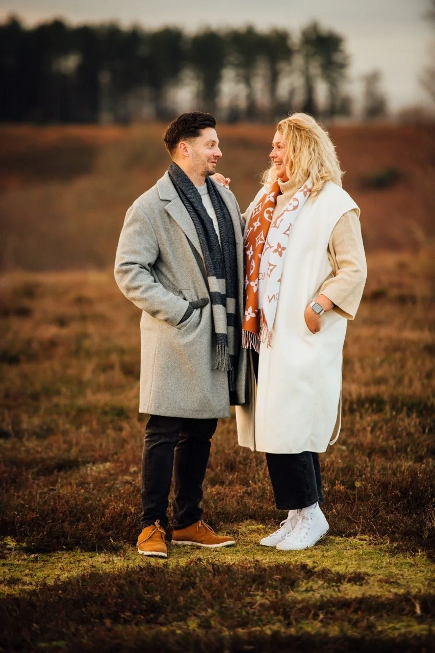 A man and a woman standing outdoors in a field, facing each other and smiling. The man has dark hair and is wearing a gray coat and a striped scarf. The woman has blonde curly hair and is wearing a white coat, a patterned scarf, and white sneakers.