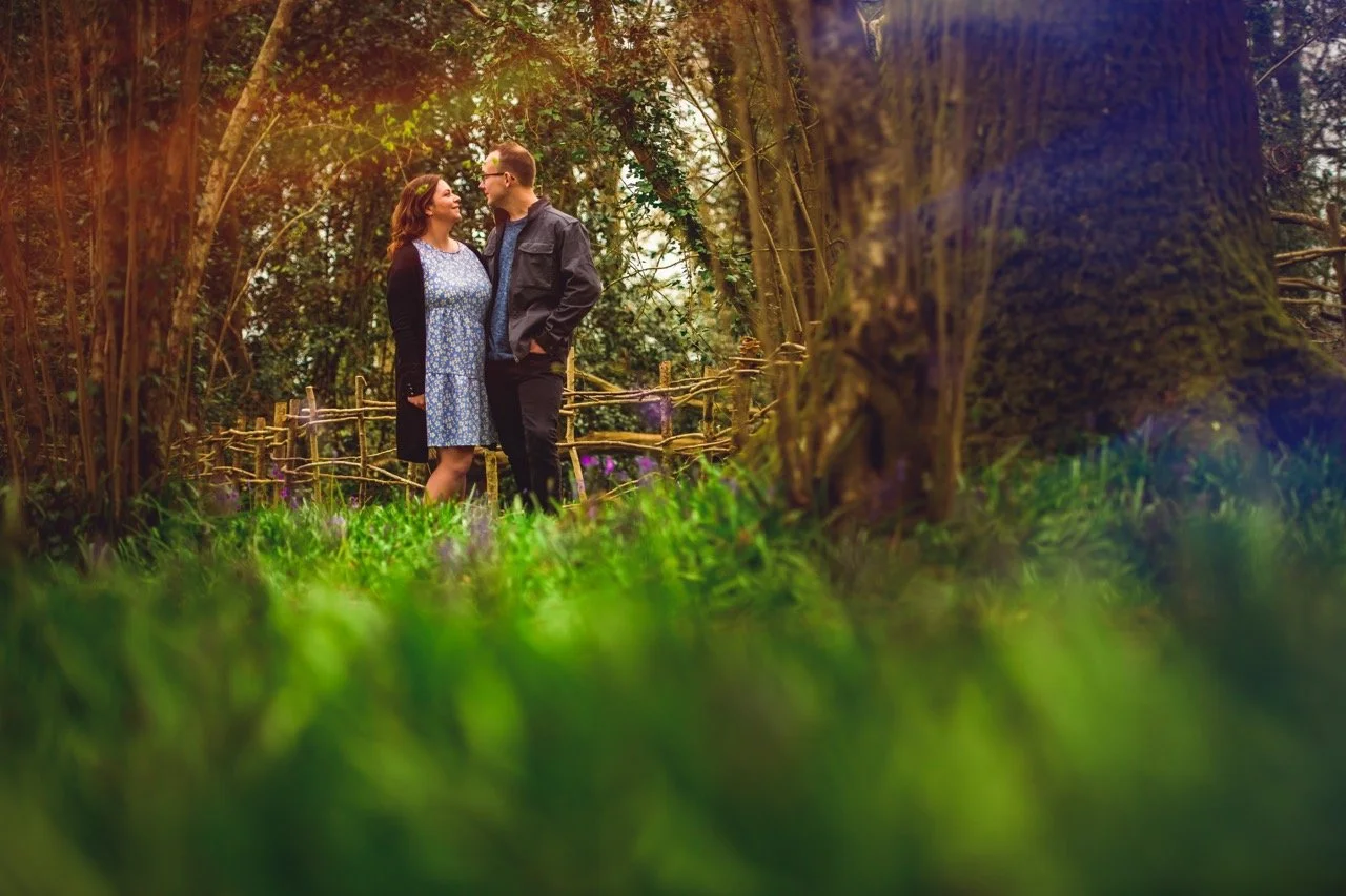 A couple standing together in a forested area, looking at each other affectionately, surrounded by trees and greenery.