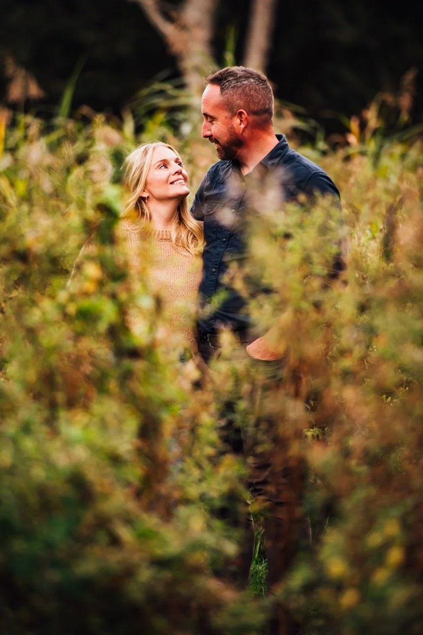 A man and woman smiling and looking at each other in a lush outdoor setting with greenery and trees.