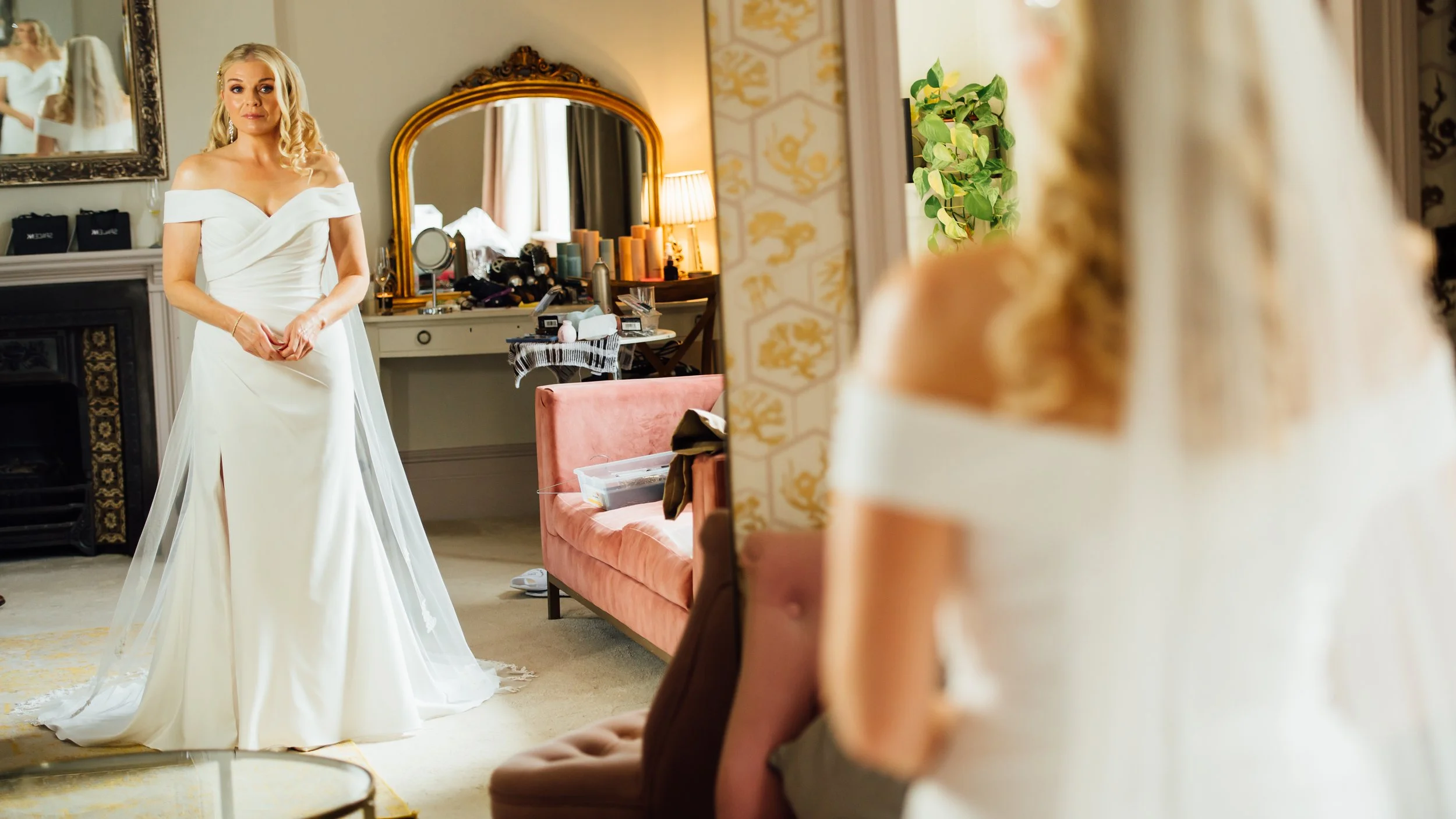 A woman in a wedding dress standing in a room, looking at her reflection in a mirror, with a woman sitting on a chair in the foreground.