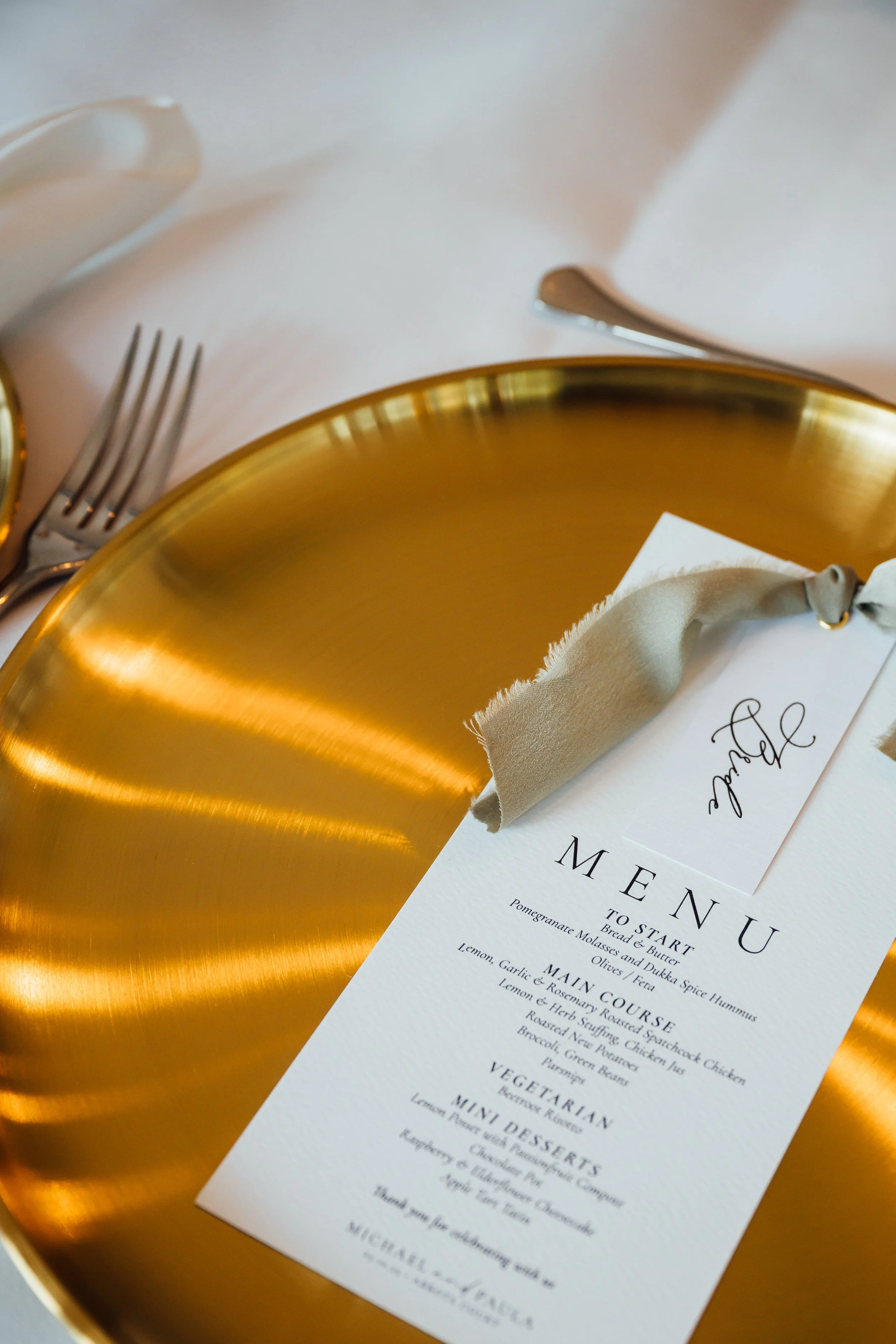 Elegant place setting with a shiny gold plate, silver fork and knife, and a white cloth napkin holding a menu card with a decorative tag.