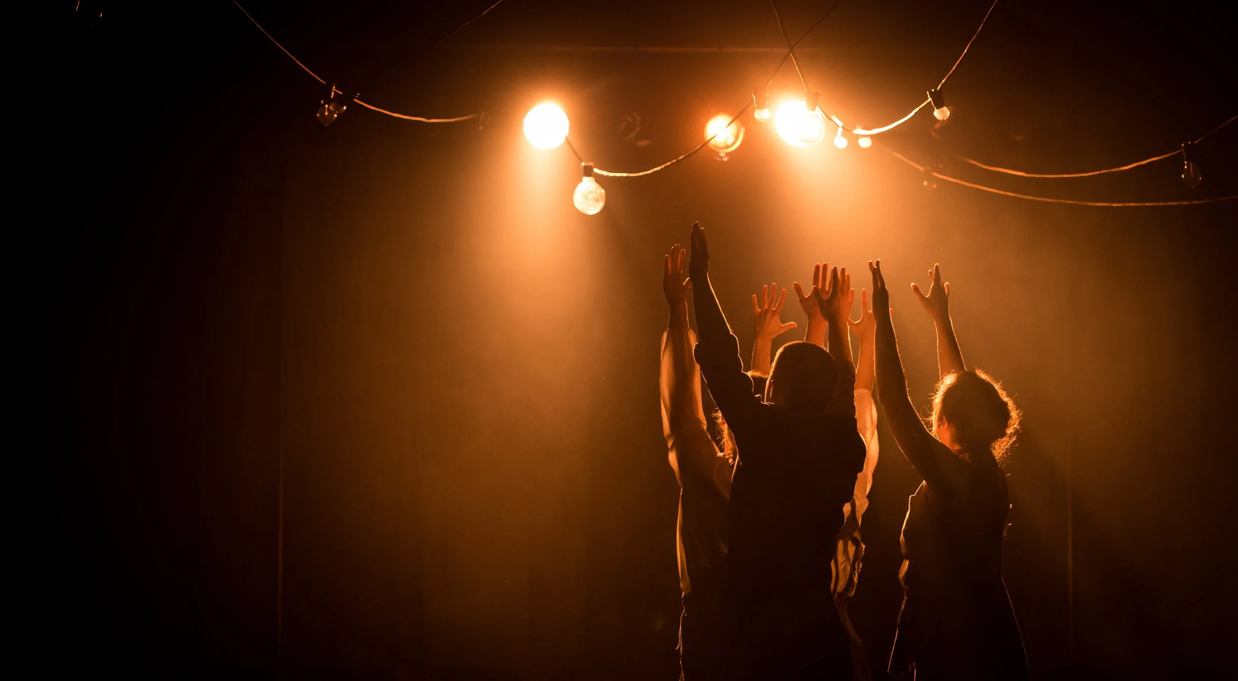 Group of 4 people in a dark stage reach with both hands towards ceiling mounted stage lights and string lights.