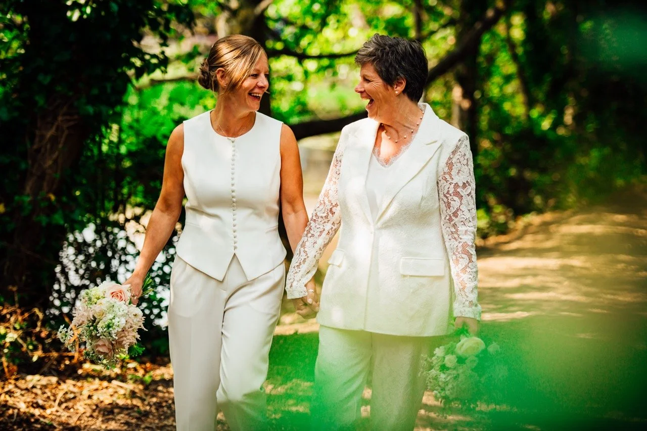 Two women in white clothing, one with a floral bouquet, walking hand in hand outdoors on a sunny day with green trees in the background, smiling and laughing.